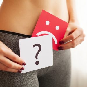 Szomorú fejet és kérdőjelet ábrázoló kártyákat tartó nő, mely az enzimhiány miatti emésztőrendszeri diszkomfort érzésre utal.